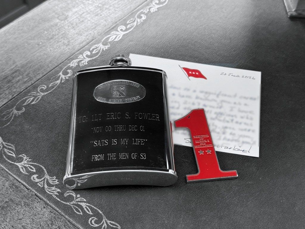 An engraved flask reading "To: 1LT Eric S. Fowler, Nov 00 thru Dec 01, SATS IS MY LIFE, from the men of S3," a red 1st Infantry Division challenge coin, and a handwritten note on three-star stationery dated 20 June 2026.